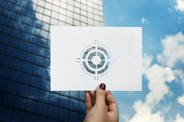 Mano sosteniendo hoja de papel con diana en stencil sobre fondo de edificio y cielo. Imagen: Teddy en Rawpixel